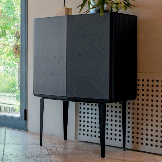 The Modern Ash Veneer Bar Cabinet, featuring sleek diagonal textured doors, stands stylishly in a modern room against a light-colored wall with a unique perforated design section. It's adorned with decorative items, including a small potted plant on top. Natural light pours in from a nearby window, enhancing the room's ambiance.
