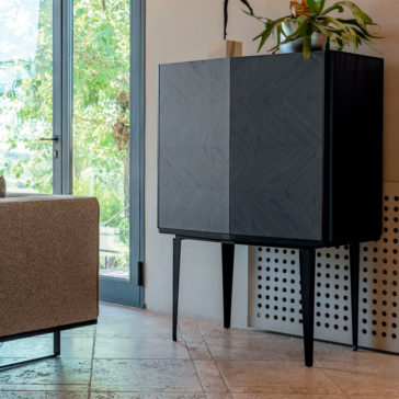 Modern Ash Veneer Bar Cabinet A Modern Ash Veneer Bar Cabinet with tall, thin legs stands in a corner next to a large glass door. The cabinet has a sleek, minimalist design and is topped with a potted plant. A beige sofa is partially visible to the left, and the floor features light-colored tiles.