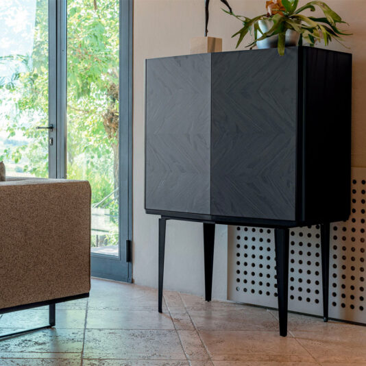 A Modern Ash Veneer Bar Cabinet with tall, thin legs stands in a corner next to a large glass door. The cabinet has a sleek, minimalist design and is topped with a potted plant. A beige sofa is partially visible to the left, and the floor features light-colored tiles.
