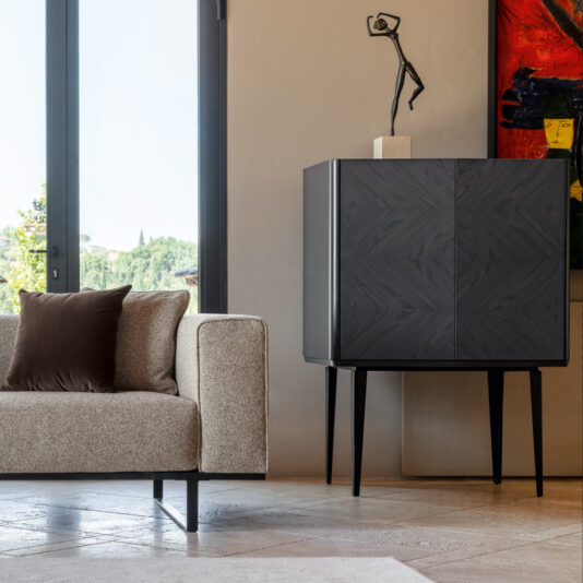 A modern living room featuring a beige sofa with a brown cushion, positioned next to the sleek Modern Ash Veneer Bar Cabinet on tall legs. The cabinet is topped with an abstract sculpture. A large window and a colorful abstract painting are also visible.