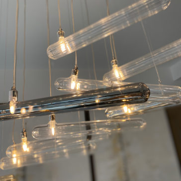 The Modern Glass Lozenge Spiral Chandelier showcases an array of elongated bulbs suspended at varying heights, each bulb casting a warm, gentle glow that crafts an elegant and contemporary ambiance against a subtly blurred backdrop.