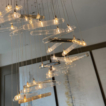 The Modern Glass Lozenge Spiral Chandelier, featuring multiple elongated light fixtures, elegantly hangs from the ceiling by thin wires. The background showcases a portion of abstract, muted art on the wall, blending neutral and soft tones.