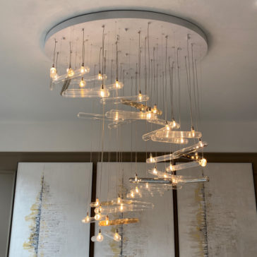 The Modern Glass Lozenge Spiral Chandelier, featuring multiple hanging lightbulbs arranged to create a mesmerizing lozenge effect, is elegantly suspended from a circular ceiling mount. In the background, abstract artwork with vertical streaks of gold and grey adorns white canvases.