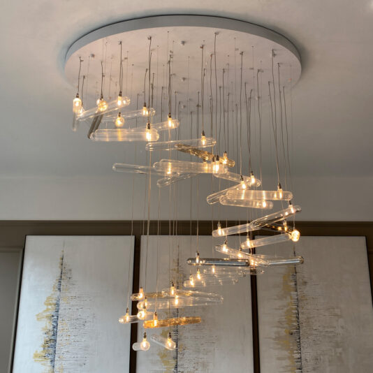 The Modern Glass Lozenge Spiral Chandelier, featuring multiple hanging lightbulbs arranged to create a mesmerizing lozenge effect, is elegantly suspended from a circular ceiling mount. In the background, abstract artwork with vertical streaks of gold and grey adorns white canvases.