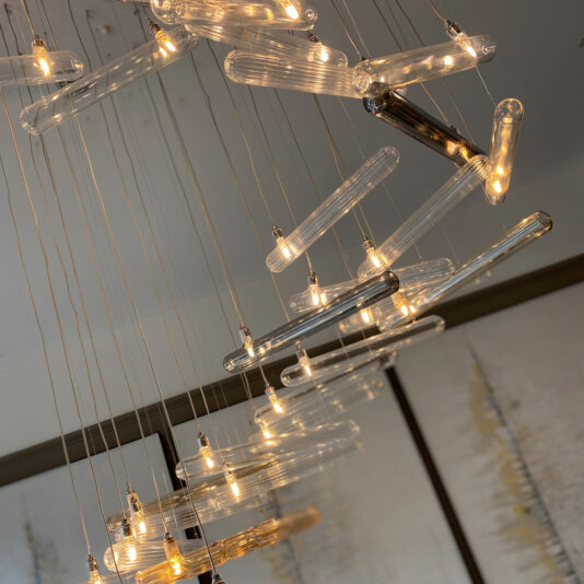 The Modern Glass Lozenge Spiral Chandelier features multiple glass rods with small internal light bulbs, suspended gracefully from the ceiling by thin wires. The glass rods are meticulously arranged in a spiral pattern, resulting in a dynamic and elegant lighting fixture.