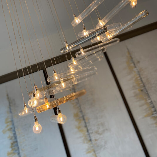 A cluster of hanging light fixtures with exposed, Edison-style light bulbs and elongated glass pendants is suspended from the ceiling, resembling the Modern Glass Lozenge Spiral Chandelier. The background features a contemporary abstract painting with vertical lines in subtle tones of yellow, white, and gray.