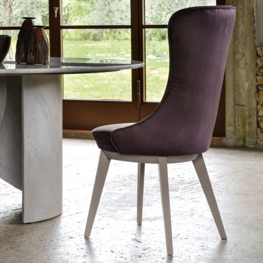 A modern dining room showcases a stylish Modern Italian Dining Chair, featuring sleek purple upholstery and white legs. The chair is positioned next to a round gray stone table, and situated near a large window that frames a scenic view of greenery and a sunlit yard beyond.