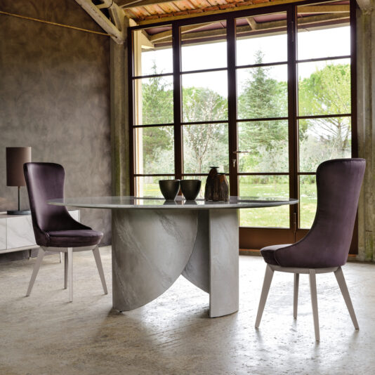 A modern, minimalistic dining space showcases a round marble table with a geometric base, flanked by two Modern Italian Dining Chairs in a muted purple tone. Large windows offer a serene view of an outdoor garden with green trees, while natural light fills the room.
