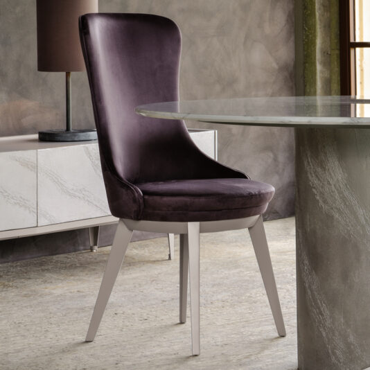In a modern dining room, there's a sleek Modern Italian Dining Chair in purple velvet with silver legs placed next to a round glass table. In the background, you can see a contemporary sideboard and a table lamp with a brown shade. The floor has a concrete finish.