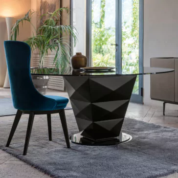 Modern Italian Glass Dining Set A contemporary dining area showcases a sleek round glass-top table from the Modern Italian Glass Dining Set, which includes a distinctive black geometric base. Accompanying the table is a plush high-backed chair upholstered in rich teal velvet. The space is adorned with green plants and large windows that let in plenty of natural light.
