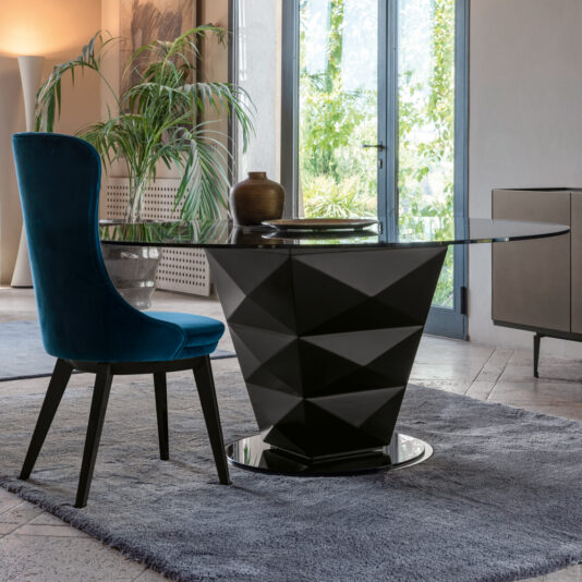 A contemporary dining area showcases a sleek round glass-top table from the Modern Italian Glass Dining Set, which includes a distinctive black geometric base. Accompanying the table is a plush high-backed chair upholstered in rich teal velvet. The space is adorned with green plants and large windows that let in plenty of natural light.