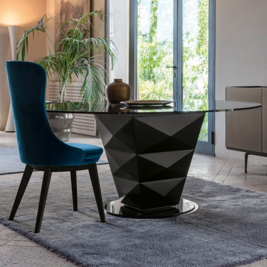 A contemporary dining area showcases a sleek round glass-top table from the Modern Italian Glass Dining Set, which includes a distinctive black geometric base. Accompanying the table is a plush high-backed chair upholstered in rich teal velvet. The space is adorned with green plants and large windows that let in plenty of natural light.