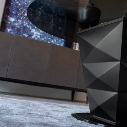 A close-up of the sleek, geometric black base of the Modern Italian Glass Dining Set highlights its exquisite design with a glass tabletop above it. In the background, a wooden cabinet stands beneath a large, colorful abstract painting adorning the wall. The floor features a light gray rug.
