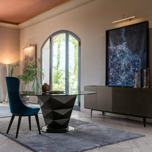 A modern dining room showcases the Modern Italian Glass Dining Set, featuring a geometric black table with a glass top and blue upholstered chairs. A large painting is displayed above a sleek grey sideboard, while a lush plant enhances the space near an arched window with doors that open to a garden view outside.