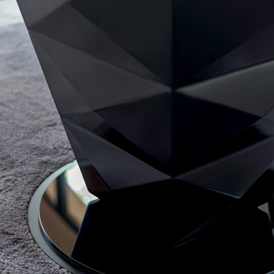 Close-up of a faceted black sculpture with a geometric design placed on a mirrored circular base. The surface around the base is covered with a light gray carpet, adjacent to the chic Modern Italian Glass Dining Set. The reflection on the mirror shows part of the room's window.