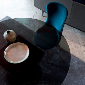 Modern Italian Glass Dining Set A teal upholstered chair sits at a round glass table, which is part of the Modern Italian Glass Dining Set, adjacent to a ceramic vase and a textured metallic plate. The arrangement features a tiled floor with a black carpet underneath the table. In the background, a dark cabinet is partially visible.