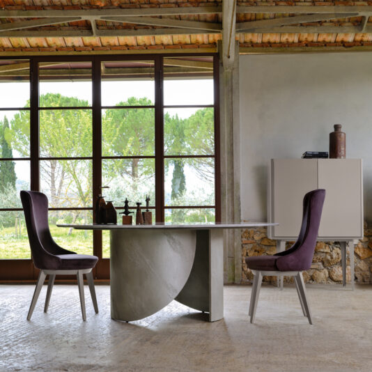 A contemporary dining room showcases a chic Modern Italian Marble Dining Set, centered by a round marble table accompanied by two elegantly upholstered purple chairs. Expansive windows offer scenic views of the surrounding trees. A beige cabinet along the wall displays various decorative pieces, and the space is enriched with ceiling beams and a textured stone wall.