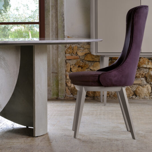 A modern dining room showcasing the Modern Italian Marble Dining Set, which includes a sleek, light grey table with a unique curved base. Accompanying it is a stylish high-back chair in deep purple velvet upholstery with light grey legs. This elegant set complements the stone wall and large window that offers a scenic view of greenery.