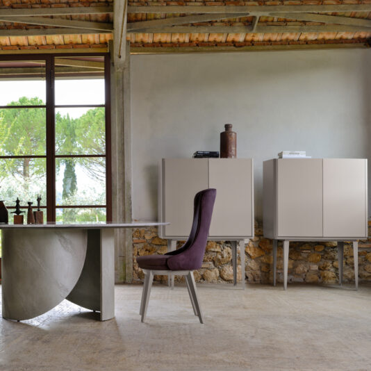 A modern interior with a large window showcases a textured table with a curved base, a purple upholstered chair, and two beige cabinets adorned with decorative items. Adding sophistication to the space is the sleek Modern Lacquered Bar Cabinet, while the ample window frames views of greenery, infusing natural light into the room.