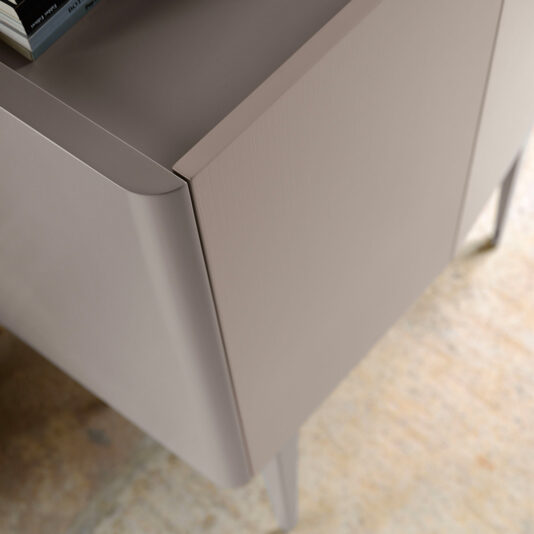 Close-up view of the corner of a modern, light beige desk with a smooth finish and minimalistic design, showing part of its top surface and leg on a light-colored, textured floor. A few books are placed on the top left corner, subtly complementing the sleek aesthetic akin to that of the Modern Lacquered Bar Cabinet.