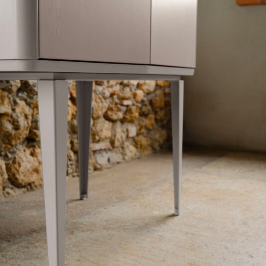 Close-up of the lower section of the Modern Lacquered Bar Cabinet, featuring thin, tapered legs. The background showcases a rustic, textured stone wall and a flat, cement-like floor, creating a striking contrast between contemporary and vintage design elements.