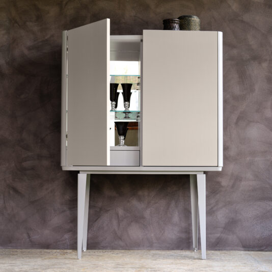 The Modern Lacquered Bar Cabinet, featuring a sleek, modern design and a matte finish, stands on tall, slender legs against a textured wall. The partially open cabinet doors reveal shelves stocked with glassware and decorative items. A few woven baskets sit atop the cabinet.