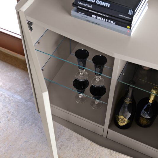 A gray Modern Lacquered Bar Cabinet with glass shelves contains black-stemmed wine glasses and wine bottles. The top of the cabinet holds three stacked books, including 