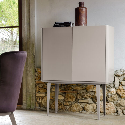 A Modern Lacquered Bar Cabinet with sleek, smooth doors stands on slim metal legs in front of a stone wall with a window beside it. A dark, cushioned chair is partially visible in the foreground. Two books and a large cylindrical object are on top of the cabinet.