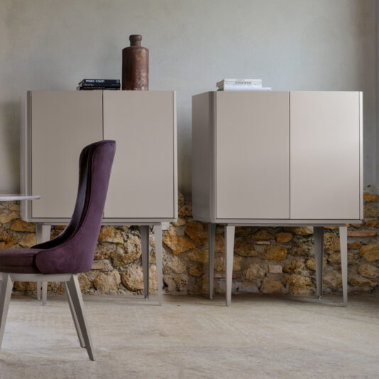 A minimalist room featuring two Modern Lacquered Bar Cabinets in light beige, each with four legs, positioned against a textured stone wall. A partially visible dark purple upholstered dining chair peeks from the left. Decorative items adorn the tops of the cabinets.