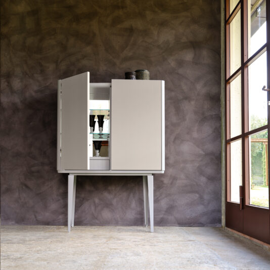 The Modern Lacquered Bar Cabinet, featuring slim, tall legs, stands poised against a textured gray wall. The partially open doors display an array of elegant tableware within. Natural light cascades through a large window on the right, beautifully illuminating the room.