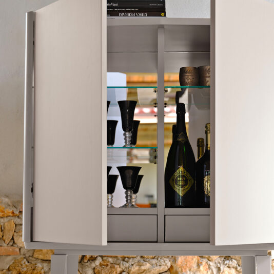 The Modern Lacquered Bar Cabinet, showcasing a sophisticated design with glass shelves, features partially open doors that reveal several black wine glasses, two unopened bottles of champagne, and two ceramic cups inside. This sleek piece stands elegantly against a rustic stone and plaster wall.