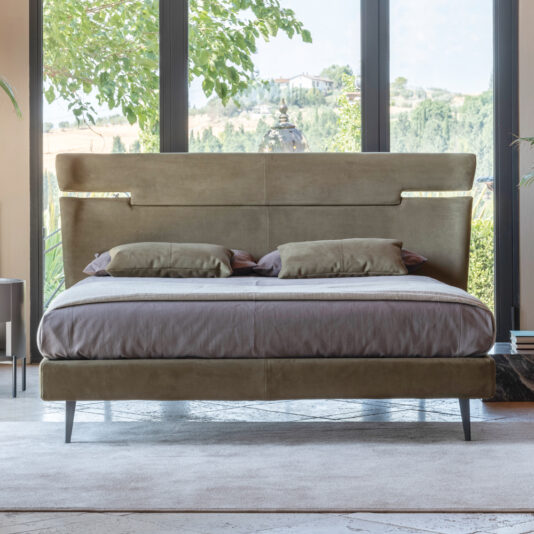 A modern bedroom showcasing the Modern Upholstered Velvet Bed, notable for its generous size, beige velvet upholstery, cushioned headboard, and matching pillows. Situated in front of expansive floor-to-ceiling windows, this bed presents a picturesque view of the surrounding greenery and rolling hills, all illuminated by abundant natural light.
