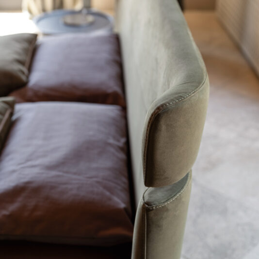Modern Upholstered Velvet Bed A close-up view of a green cushioned sofa with brown pillows. The image, taken from a high angle, showcases the armrest and backrest in focus. A side table is visible in the background, enhancing the charm of the well-lit indoor space. This elegant setting is perfectly complemented by the Modern Upholstered Velvet Bed.
