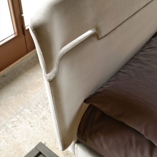 Modern Upholstered Velvet Bed A close-up view of the Modern Upholstered Velvet Bed showcases its stylish design with a light grey, upholstered velvet headboard. Positioned beside a large window, the dark bedding partially visible adds a striking contrast to the lighter headboard and surrounding decor.