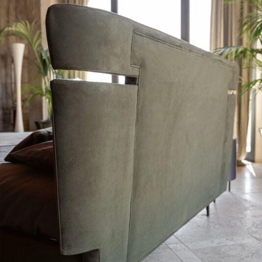 Modern Upholstered Velvet Bed A close-up view of a stylish green sofa featuring a distinctive armrest design in an airy room. Positioned near expansive windows, the sofa is bathed in natural light that illuminates the room's verdant plants and chic decor elements, evoking the elegance of a Modern Upholstered Velvet Bed.