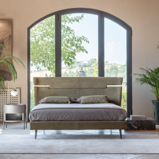 Modern Upholstered Velvet Bed A modern bedroom showcases the Modern Upholstered Velvet Bed, sleek and beige, adorned with pillows and a throw blanket. This elegant bed is placed in front of a large arched window offering a serene view of the greenery outside. To the left, a small bedside table with a lamp provides additional functionality, while large potted plants further enhance the decor.
