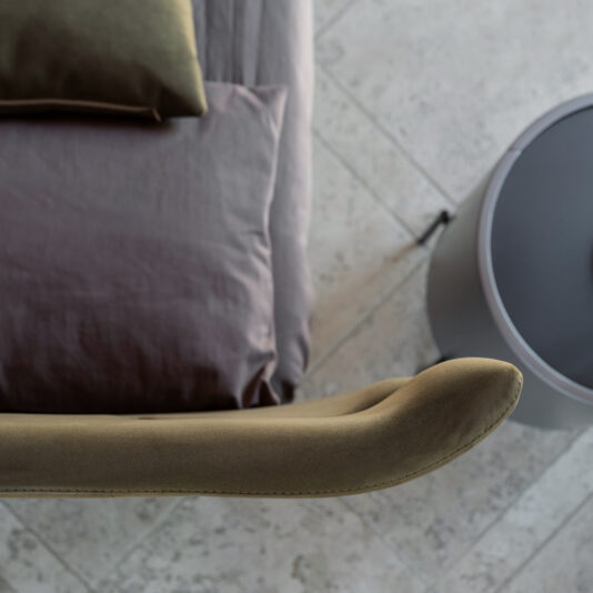 Modern Upholstered Velvet Bed Top-down view of a minimalist bedroom corner featuring the Modern Upholstered Velvet Bed with green and gray bedding, a cylindrical nightstand, and part of a green padded chair. The room has a tiled floor with a textured design.