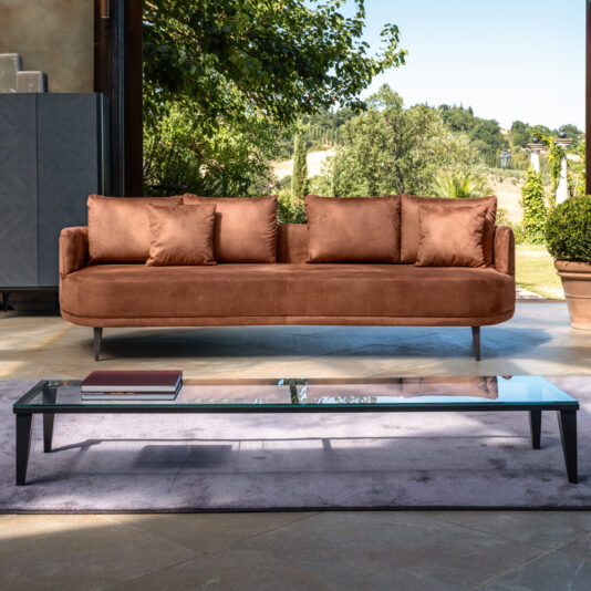 A sleek, modern living room showcasing a burnt orange sofa with clean lines and cushions sits in front of large windows that display a lush, green outdoor landscape. A Narrow Glass Coffee Table stands on a grey area rug in front of the sofa, holding a few books.