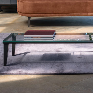 Narrow Glass Coffee Table A Narrow Glass Coffee Table with black legs, holding a stack of two hardcover books, sits on a light purple rug. In the background, there is a portion of a brown upholstered sofa on a beige tiled floor. Bright natural light illuminates the scene.