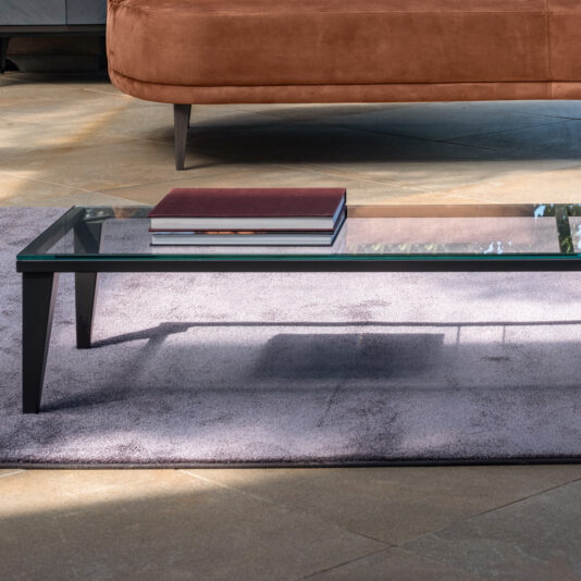 A Narrow Glass Coffee Table with black legs, holding a stack of two hardcover books, sits on a light purple rug. In the background, there is a portion of a brown upholstered sofa on a beige tiled floor. Bright natural light illuminates the scene.