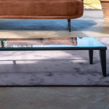 Narrow Glass Coffee Table A modern living room scene featuring the Narrow Glass Coffee Table with black legs on a grayish-purple rug. In the background, a rust-colored sofa with wooden legs sits on a light gray tile floor. Potted plants are visible outside through a glass window.