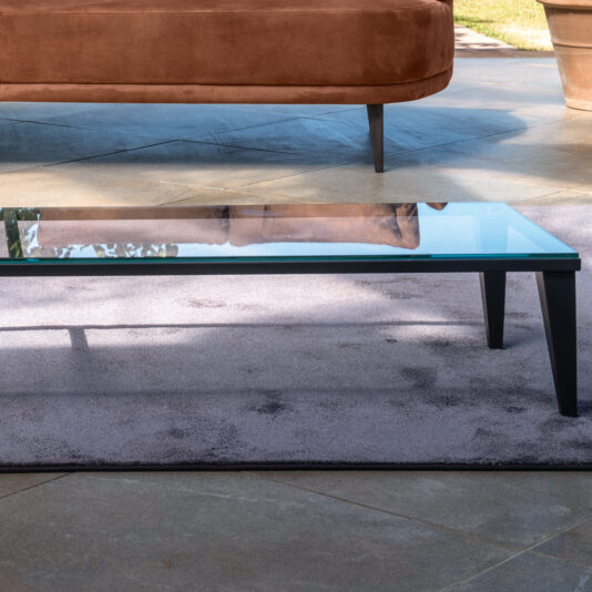 A modern living room scene featuring the Narrow Glass Coffee Table with black legs on a grayish-purple rug. In the background, a rust-colored sofa with wooden legs sits on a light gray tile floor. Potted plants are visible outside through a glass window.