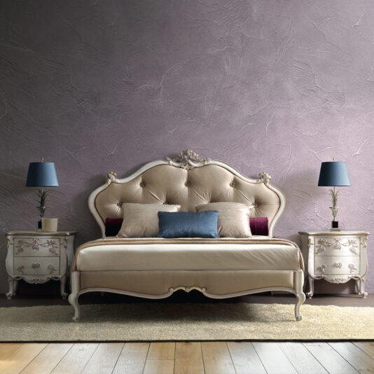 Rococo Style Button Upholstered Bed A bedroom features a sophisticated Rococo Style Button Upholstered Bed with beige fabric and multiple pillows in neutral tones. Flanking the bed are two ornate white nightstands, each adorned with a navy blue lamp. The wall behind the bed is textured and painted in a muted lavender hue.
