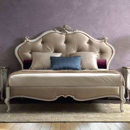The Rococo Style Button Upholstered Bed features a luxurious tufted headboard with ornate, intricate details and a lavish design. Adorned with beige bedding, two large beige pillows, and a blue accent pillow at the front, this bed is set against a textured purple wall and stands elegantly on a light wooden floor.