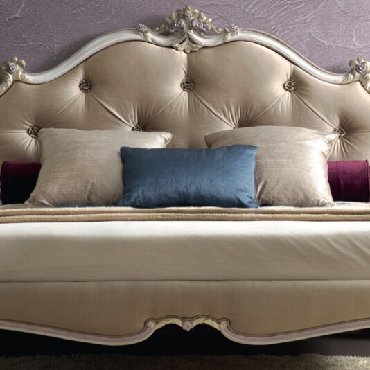 Rococo Style Button Upholstered Bed The Rococo Style Button Upholstered Bed features an elegant, tufted headboard with carved wooden details in a creamy beige tone. It is adorned with plush pillows in shades of beige, dark blue, and burgundy, creating a luxurious bed setup that is perfectly complemented by a textured lavender wall in the background.