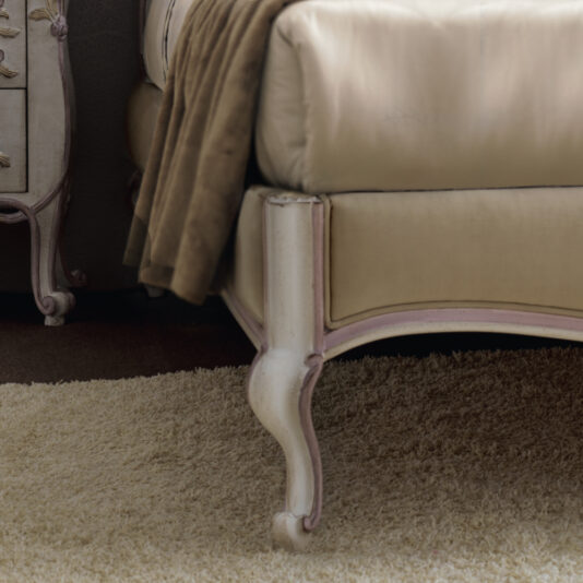 Rococo Style Button Upholstered Bed A close-up shot highlights the exquisitely designed Rococo Style Button Upholstered Bed, featuring its cream-colored upholstery and ornate curved legs set against a light beige carpet. A brown throw is casually draped over the bed, while part of a matching ornate bedside table peeks into view in the background.