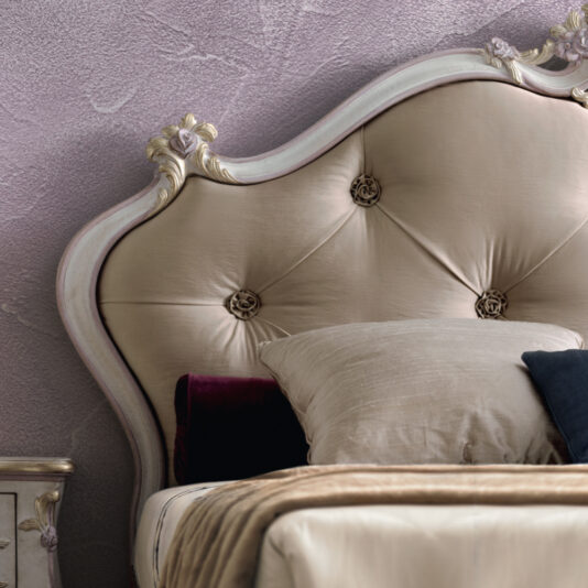 Rococo Style Button Upholstered Bed A close-up of the Rococo Style Button Upholstered Bed showcases an elegant headboard with intricate floral details in a soft beige color. The bed is adorned with neatly arranged pillows in various shades of beige, black, and deep purple, set against a textured light purple wall.