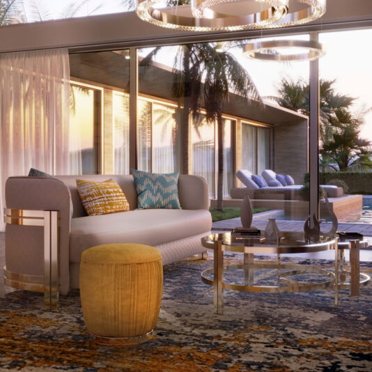 A modern living room with a large glass wall showcasing a sunset view. The room features a light-colored sofa adorned with patterned throw pillows, accompanied by a round glass coffee table. Beside the yellow ottoman on the patterned rug stands the Ruched Pouffe With Gold Plated Base. Outside, lounge chairs and lush palm trees complete the scene.
