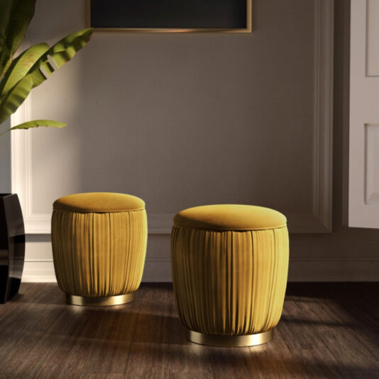 Two Ruched Pouffes with Gold Plated Bases in yellow sit gracefully on a dark wooden floor. A large green plant and part of a white wall and door are visible in the background, suggesting the scene is in a modern, stylish indoor space.