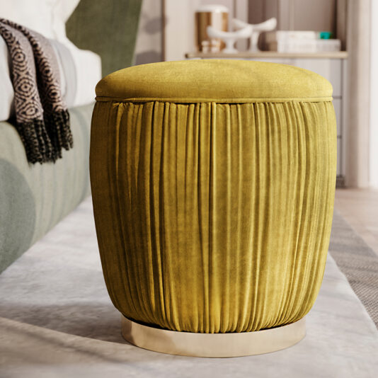 A Ruched Pouffe With Gold Plated Base, in a mustard yellow color, sits on a faintly patterned rug. Behind it, a green fabric sofa with a black and white patterned blanket draped over the arm is partially visible. The background includes a neutral-colored room.